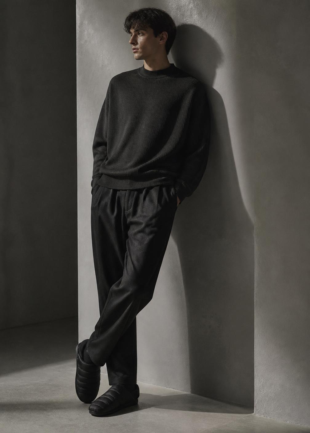 Black padded slippers styled with dark tailoring against a plaster wall